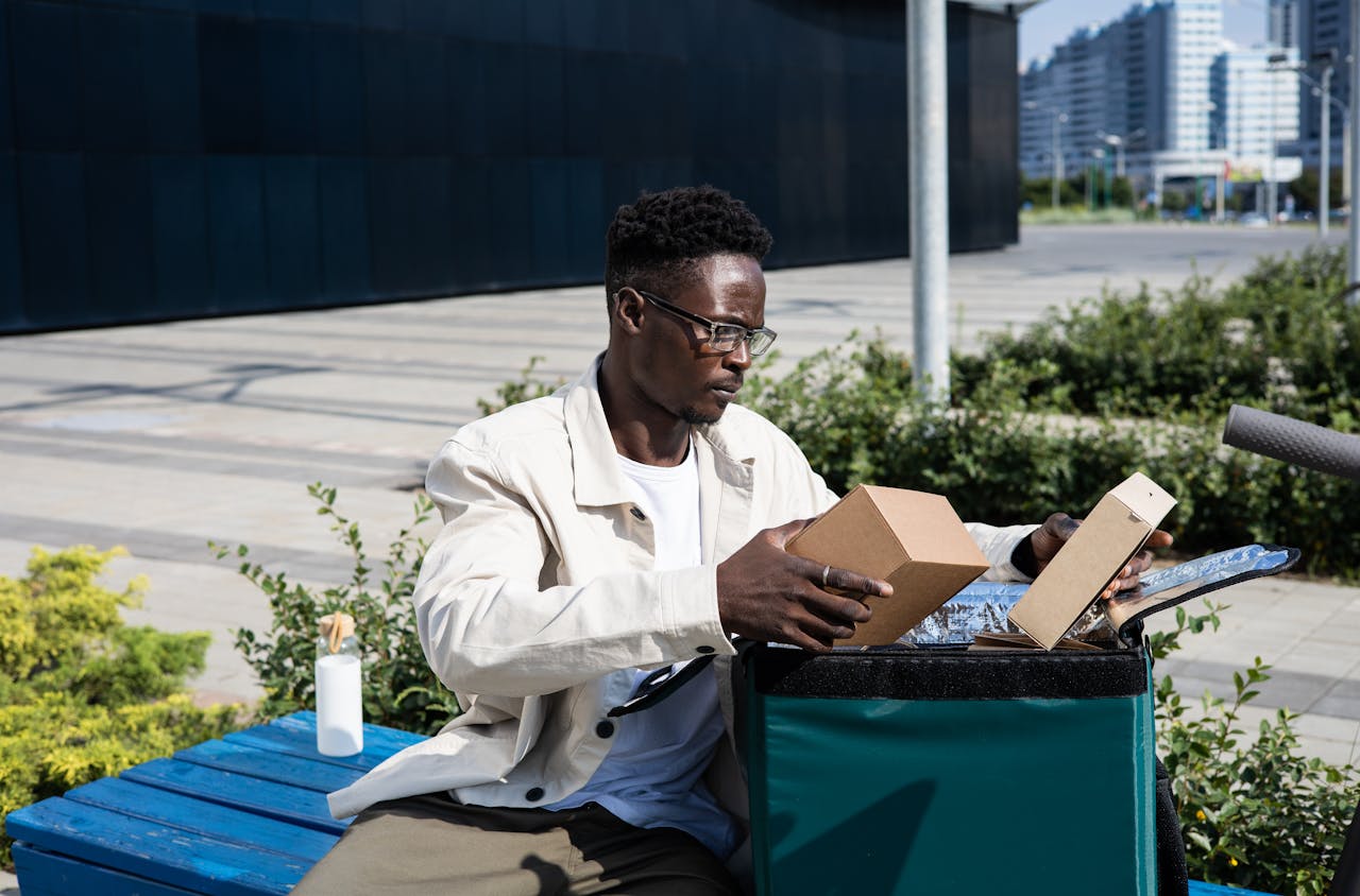A delivery man sitting outdoors organizing parcels from a thermal bag on a bench.